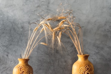 Two ornately decorated ceramic vases are positioned side by side against a textured gray background. Each vase holds golden stalks of wheat that curve gracefully, creating an arch above the vases. The pottery features artistic designs etched into its surface, adding to the rustic and natural aesthetic.