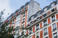A multi-story residential building features a classic architectural design with white and orange facade accents and wrought-iron railings on the balconies. The structure includes numerous windows and a steep, dark roof. Some greenery, possibly from nearby trees, partially obscures the lower part of the building.