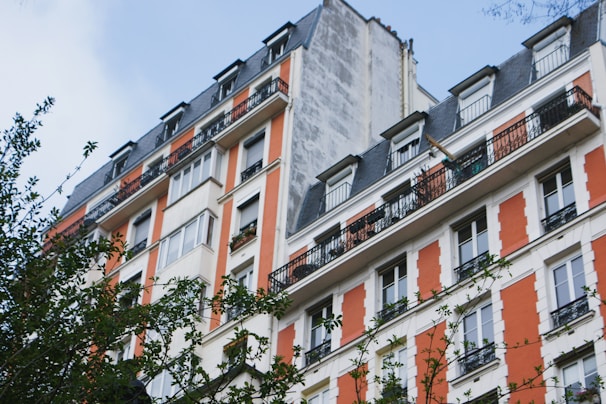 A multi-story residential building features a classic architectural design with white and orange facade accents and wrought-iron railings on the balconies. The structure includes numerous windows and a steep, dark roof. Some greenery, possibly from nearby trees, partially obscures the lower part of the building.