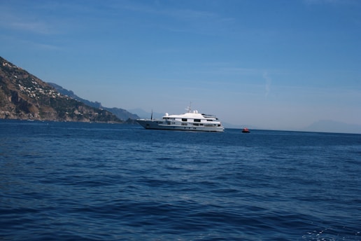 A large, white yacht is sailing on a calm blue sea, with a distant coastline featuring rugged hills and sparse vegetation. The sky is clear and bright, indicating a sunny day.