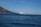 A large, white yacht is sailing on a calm blue sea, with a distant coastline featuring rugged hills and sparse vegetation. The sky is clear and bright, indicating a sunny day.