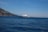 A large, white yacht is sailing on a calm blue sea, with a distant coastline featuring rugged hills and sparse vegetation. The sky is clear and bright, indicating a sunny day.