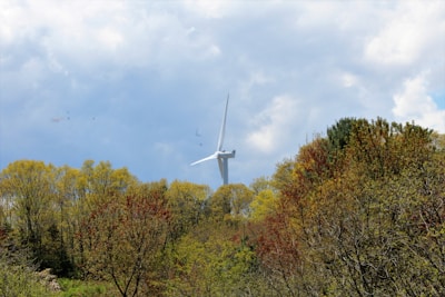 Wind turbine standing tall amidst a lush forest, with its blades extending into a partly cloudy sky. The trees exhibit varying shades of green, yellow, and hints of red, indicating a transition period in nature. The scene is tranquil, highlighting both technological advancement and natural beauty.
