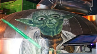 A large mural depicts a green humanoid figure with pointed ears and white hair, wearing a robe and holding a glowing green lightsaber. The setting includes industrial elements like metal ducts and a control panel screen nearby.