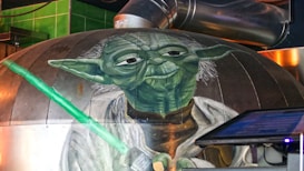 A large mural depicts a green humanoid figure with pointed ears and white hair, wearing a robe and holding a glowing green lightsaber. The setting includes industrial elements like metal ducts and a control panel screen nearby.