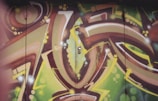 An abstract graffiti artwork featuring curved lines and geometric shapes in vibrant colors. The design includes gradients of green, brown, yellow, and white, with spots and swirls adding depth to the piece. The composition is dynamic, with a sense of movement and flow.