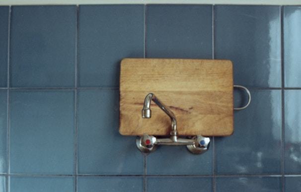 A wooden cutting board is attached to a wall, with a metal faucet extending outward. The background consists of blue ceramic tiles, giving a clean and minimalist look.