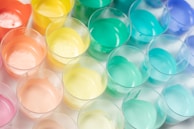 An array of plastic cups filled with pastel-colored paints arranged closely together. The colors range from orange, yellow, mint green to blue, creating a visually appealing pattern. The surface of each paint appears smooth and glossy under the light.