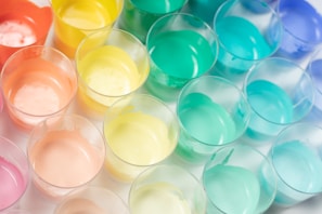An array of plastic cups filled with pastel-colored paints arranged closely together. The colors range from orange, yellow, mint green to blue, creating a visually appealing pattern. The surface of each paint appears smooth and glossy under the light.