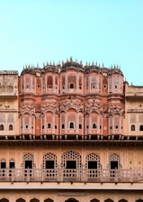 An ornate architectural facade featuring intricate latticework and multiple small windows. The structure has a series of symmetrical archways and decorative elements typical of traditional Indian design. The building displays various shades of pink and beige, set against a clear blue sky.
