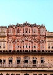 An ornate architectural facade featuring intricate latticework and multiple small windows. The structure has a series of symmetrical archways and decorative elements typical of traditional Indian design. The building displays various shades of pink and beige, set against a clear blue sky.