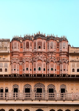 An ornate architectural facade featuring intricate latticework and multiple small windows. The structure has a series of symmetrical archways and decorative elements typical of traditional Indian design. The building displays various shades of pink and beige, set against a clear blue sky.
