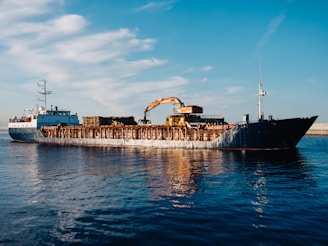 A large cargo ship is sailing on a calm body of water. The ship appears to be carrying heavy machinery and equipment, including an industrial crane on its deck. The sky is clear with a few scattered clouds, creating a serene backdrop to the industrial scene. The water reflects the blue sky and the ship, adding to the tranquility of the setting.