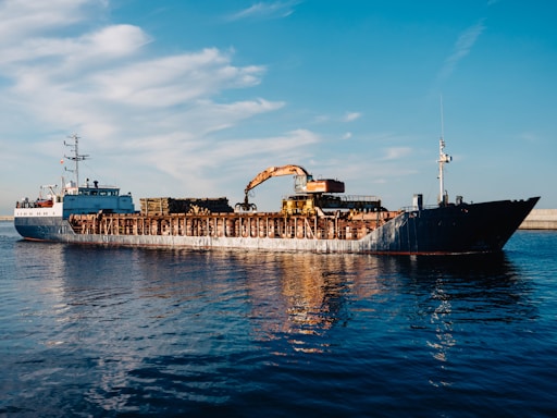 A large cargo ship is sailing on a calm body of water. The ship appears to be carrying heavy machinery and equipment, including an industrial crane on its deck. The sky is clear with a few scattered clouds, creating a serene backdrop to the industrial scene. The water reflects the blue sky and the ship, adding to the tranquility of the setting.