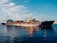 A large cargo ship is sailing on a calm body of water. The ship appears to be carrying heavy machinery and equipment, including an industrial crane on its deck. The sky is clear with a few scattered clouds, creating a serene backdrop to the industrial scene. The water reflects the blue sky and the ship, adding to the tranquility of the setting.