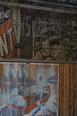 The image features a vintage mural on a brick wall with an old advertisement. One portion shows a faded advertisement with a person depicted in a black and white style, while another portion shows a colorful illustration of two people in a canoe. The canoe image is painted on wooden panels with a worn texture, using shades of blue, orange, and white.