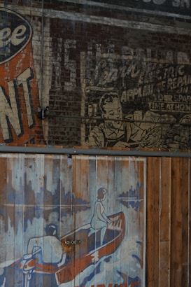 The image features a vintage mural on a brick wall with an old advertisement. One portion shows a faded advertisement with a person depicted in a black and white style, while another portion shows a colorful illustration of two people in a canoe. The canoe image is painted on wooden panels with a worn texture, using shades of blue, orange, and white.