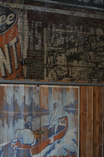 The image features a vintage mural on a brick wall with an old advertisement. One portion shows a faded advertisement with a person depicted in a black and white style, while another portion shows a colorful illustration of two people in a canoe. The canoe image is painted on wooden panels with a worn texture, using shades of blue, orange, and white.