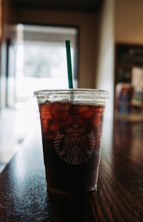 A plastic cup filled with iced coffee featuring a logo is positioned on a wooden surface, with a green straw emerging from the top. The background is softly blurred and suggests an indoor setting with ambient light coming through a window.