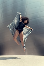 A dancer with curly hair is captured mid-air, wearing a black outfit and a flowing, sheer fabric that accentuates their movement. The background features a large, metallic wall with a modern architectural design, while the ground is a smooth, light-colored surface.