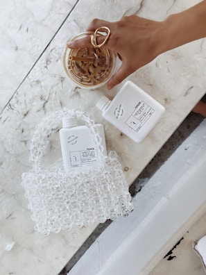 A hand adorned with a large, gold ring is holding a cup of iced coffee on a marble surface. Beside it, there is a transparent beaded handbag and two identical white bottles labeled 'TONIK'. The scene is minimalist with a focus on the products and accessories.