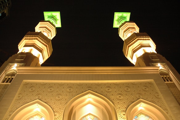 A night view of a building with two illuminated minarets featuring intricate architectural designs. The tops of the minarets are glowing green, contributing to the dramatic night ambiance. The facade displays detailed patterns and arches that are highlighted by warm lighting.