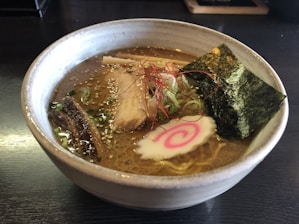 A bowl of ramen with rich broth, topped with slices of pork, seaweed, narutomaki, green onions, and thin strands of red chili pepper.