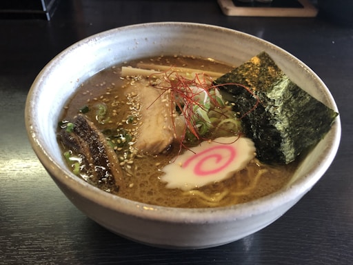 A bowl of ramen with rich broth, topped with slices of pork, seaweed, narutomaki, green onions, and thin strands of red chili pepper.