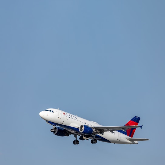 A passenger airplane with the Delta logo is captured in flight against a clear blue sky. The aircraft, identifiable by its white fuselage and prominent red and blue markings on the tail, appears to be ascending.