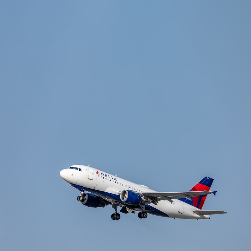 A passenger airplane with the Delta logo is captured in flight against a clear blue sky. The aircraft, identifiable by its white fuselage and prominent red and blue markings on the tail, appears to be ascending.