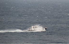A small pilot boat navigates through a large expanse of ocean, creating a trail of white spray as it cuts through the choppy, dark blue waters. The words 'PILOT' are visible on the side of the boat.
