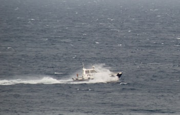 A small pilot boat navigates through a large expanse of ocean, creating a trail of white spray as it cuts through the choppy, dark blue waters. The words 'PILOT' are visible on the side of the boat.