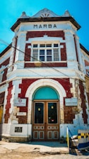 A historic building with a combination of red bricks and white accents, featuring a central arched doorway with teal highlights. Various signs, including 'Marba' at the top, are visible. The structure has a classical architectural style with decorative elements around the roofline.