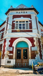 A historic building with a combination of red bricks and white accents, featuring a central arched doorway with teal highlights. Various signs, including 'Marba' at the top, are visible. The structure has a classical architectural style with decorative elements around the roofline.