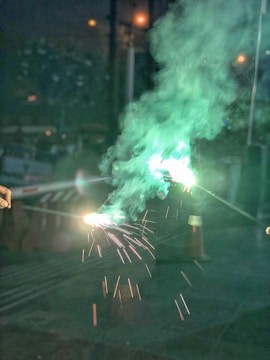Two sparklers are being held, emitting bright sparks and green smoke in a dimly lit environment. The background shows some faint lights and indistinct structures.