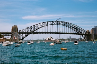 A large arch bridge spans across a body of water surrounded by several small boats. The iconic Sydney Opera House is visible in the background. The scene includes a ferris wheel and buildings to the left. The sky is clear and blue, adding to the scenic ambiance.