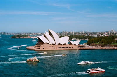 A panoramic view of the iconic Sydney Opera House surrounded by the vibrant waters of Sydney Harbour. Several boats and ferries are seen navigating the harbor, creating trails of white foam on the blue water. The background consists of lush greenery and a bustling cityscape with numerous buildings under a clear blue sky.