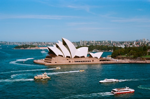 A panoramic view of the iconic Sydney Opera House surrounded by the vibrant waters of Sydney Harbour. Several boats and ferries are seen navigating the harbor, creating trails of white foam on the blue water. The background consists of lush greenery and a bustling cityscape with numerous buildings under a clear blue sky.