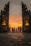 A group of people is walking towards a dramatic stone gate adorned with intricate carvings. The sky behind them is illuminated with deep oranges and yellows, suggesting a sunset. Two statues, likely of deities or mythological figures, are prominently placed on either side of the gate, adding an element of grandeur. The pavement leading to the gate is constructed of large stone tiles.