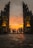 A group of people is walking towards a dramatic stone gate adorned with intricate carvings. The sky behind them is illuminated with deep oranges and yellows, suggesting a sunset. Two statues, likely of deities or mythological figures, are prominently placed on either side of the gate, adding an element of grandeur. The pavement leading to the gate is constructed of large stone tiles.