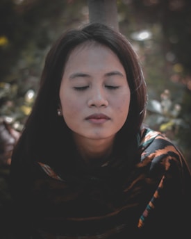 A woman is seen in a close-up portrait with her eyes closed, exuding a sense of calm and serenity. She has dark hair and is wearing earrings and a patterned garment. The background is blurred, featuring hints of greenery.