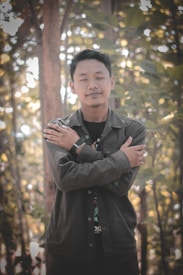 A person stands in a forest setting with eyes closed and arms crossed over their chest, conveying a peaceful or meditative pose. Sunlight filters through the leaves, casting a warm, diffused glow on the scene.