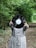 Person wearing a checkered outfit is holding a vinyl record in front of their face in a wooded area with a path. The person and the large black vinyl stand out against the lush green background of trees and bushes.