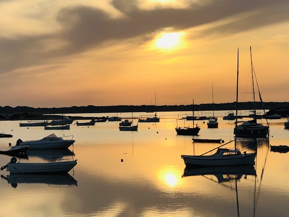 Sunset view over calm waters with sailboats anchored near the shore.