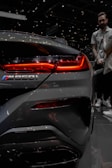 A close-up of the rear section of a sleek, metallic gray car focusing on the taillight and the M850i logo. The vibrant red light contrasts sharply against the dark and reflective surface of the vehicle. In the blurred background, a person stands at an auto show, adding an element of indoor lighting and atmosphere to the scene.