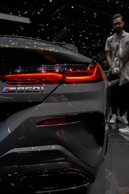 A close-up of the rear section of a sleek, metallic gray car focusing on the taillight and the M850i logo. The vibrant red light contrasts sharply against the dark and reflective surface of the vehicle. In the blurred background, a person stands at an auto show, adding an element of indoor lighting and atmosphere to the scene.