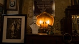 A dimly lit interior features an eclectic collection of items, including a framed portrait of a woman, a white bust sculpture of a man, a vintage lamp with exposed bulbs, and a decorative ceramic figure. A poster is mounted on a stone wall in the background, and a wooden cabinet with ornate details is visible.