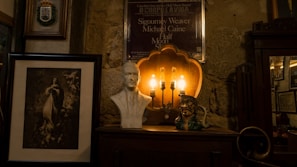 A dimly lit interior features an eclectic collection of items, including a framed portrait of a woman, a white bust sculpture of a man, a vintage lamp with exposed bulbs, and a decorative ceramic figure. A poster is mounted on a stone wall in the background, and a wooden cabinet with ornate details is visible.
