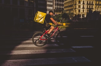 A cyclist is riding on a crosswalk in a city setting, carrying a large yellow Glovo delivery bag on a red bicycle. The environment features urban architecture with sunlight creating a strong contrast between light and shadow.