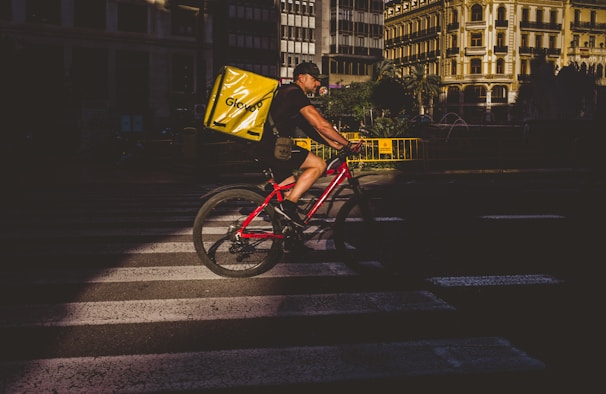 A cyclist is riding on a crosswalk in a city setting, carrying a large yellow Glovo delivery bag on a red bicycle. The environment features urban architecture with sunlight creating a strong contrast between light and shadow.